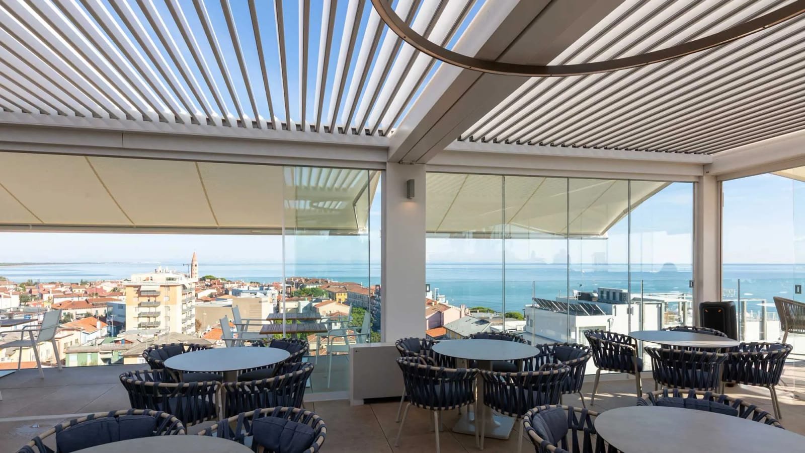 Terraza de restaurante en una azotea con pérgola bioclimática de lamas blancas y cerramiento de cristal frente al mar
